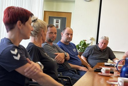 Mehrere Personen sitzen an einem langen Holztisch in einem Besprechungsraum. Auf dem Tisch stehen Tassen, Untertassen, Thermoskannen und Notizmaterial. Im Hintergrund sind ein Fenster mit vertikalen Lamellen, eine Tür, eine Zimmerpflanze und eine Projektionsfläche zu sehen.