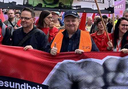 Mehrere Personen stehen dicht gedrängt hinter einem großen roten Banner mit einer schwarz-weißen Faustgrafik. In der Bildmitte trägt eine Person eine orange Warnweste mit dem Aufdruck „EVG“ und eine dunkle Kappe mit „EVG“ und zeigt einen erhobenen Daumen. Im Hintergrund sind zahlreiche rote Fahnen, weitere Personen sowie ein teilweise sichtbares Transparent mit Industrieanlagen und Windrädern abgebildet. Auf dem Banner ist der Textausschnitt „Gesundheit“ sichtbar.