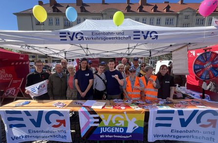 Gruppenfoto an einem Infostand im Freien: Ein weißes Pavillonzelt mit der Aufschrift „EVG Eisenbahn- und Verkehrsgewerkschaft“ steht auf einem Platz mit historischen Gebäuden im Hintergrund. Unter dem Zelt stehen mehrere Personen hinter einem langen Tisch. Auf dem Tisch liegen Flyer, Broschüren, Aufkleber und Informationsmaterial. An der Vorderseite des Tisches hängen Banner mit dem EVG-Logo sowie ein farbiges Banner mit der Aufschrift „EVG JUGEND – Wir leben Gemeinschaft“. Einige Personen tragen orangefarbene Warnwesten mit EVG-Logo, andere dunkle Oberteile mit EVG-Schriftzug. Über dem Zelt sind bunte Luftballons befestigt. Links und rechts sind weitere rote und weiße Gewerkschaftsstände mit Fahnen und einem Glücksrad sichtbar.