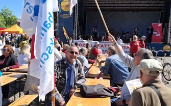 Großer Platz im Freien mit Biertischen und -bänken, vielen sitzenden und stehenden Personen. Im Vordergrund ein Tisch mit einer Person mit Sonnenbrille, Schal und Jacke. Mehrere Fahnen und Banner mit Gewerkschaftslogos sind sichtbar. Im Hintergrund eine Bühne mit Dach, Musikinstrumenten und mehreren Personen auf der Bühne. Auf einem Banner an der Bühne steht „entega“ und „EINFACH KLIMAFREUNDLICH FÜR ALLE“.