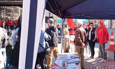 Blick durch ein blaues Pavillonzelt auf einen Infotisch mit ausliegenden Zeitungen, Broschüren und Flyern. Mehrere Personen stehen davor und daneben. Im Hintergrund sind weitere rote und blaue Pavillons, Fahnen mit EVG‑ und DGB‑Logos sowie eine Bühne mit Personen zu sehen. Der Boden ist gepflastert.