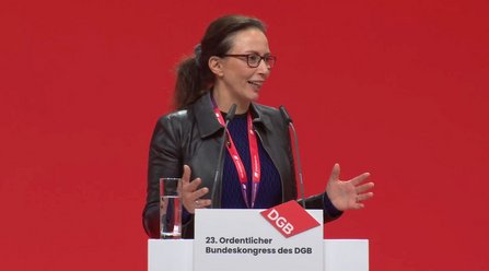 Eine Person steht hinter einem weißen Rednerpult vor rotem Hintergrund. Auf dem Pult befinden sich zwei Mikrofone und ein Glas mit Wasser. Die Person trägt eine dunkle Jacke über einem dunklen Oberteil und ein rotes Schlüsselband. Beide Hände sind seitlich vor dem Körper erhoben. Auf der Vorderseite des Rednerpults steht das Logo „DGB“ sowie der Text „23. Ordentlicher Bundeskongress des DGB“.