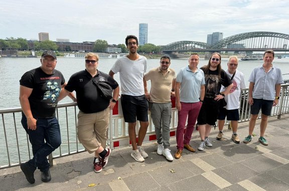 Neun Personen stehen nebeneinander an einem Geländer entlang eines Flusses. Im Hintergrund sind eine große Bogenbrücke aus Stahl, weitere Gebäude sowie ein hohes modernes Gebäude zu sehen. Auf dem Wasser fährt ein weißes Schiff. Die Personen tragen Freizeitkleidung wie T‑Shirts, Poloshirts, Shorts, lange Hosen und Turnschuhe; einige tragen Sonnenbrillen. Mehrere Hände liegen auf dem Geländer oder sind in die Hüfte gestützt. Der Himmel ist hell und leicht bewölkt, das Ufer ist gepflastert.