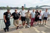 Neun Personen stehen nebeneinander an einem Geländer entlang eines Flusses. Im Hintergrund sind eine große Bogenbrücke aus Stahl, weitere Gebäude sowie ein hohes modernes Gebäude zu sehen. Auf dem Wasser fährt ein weißes Schiff. Die Personen tragen Freizeitkleidung wie T‑Shirts, Poloshirts, Shorts, lange Hosen und Turnschuhe; einige tragen Sonnenbrillen. Mehrere Hände liegen auf dem Geländer oder sind in die Hüfte gestützt. Der Himmel ist hell und leicht bewölkt, das Ufer ist gepflastert.