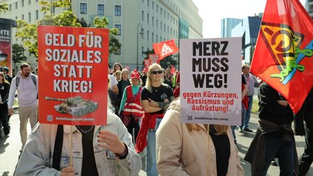 Zwei Personen tragen Schilder im Demonstrationszug. Das linke Schild trägt den Text „GELD FÜR SOZIALES STATT KRIEG!“, das rechte Schild den Text „MERZ MUSS WEG! gegen Kürzungen, Rassismus und Aufrüstung!“. Im Hintergrund sind weitere Teilnehmende, rote Fahnen und städtische Gebäude zu sehen.