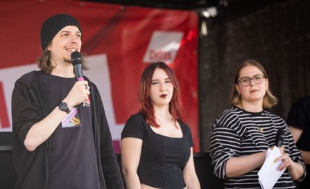 Drei Personen stehen auf einer Bühne vor einem roten Hintergrund mit teilweise sichtbarem DGB-Logo. Die linke Person trägt eine dunkle Mütze und hält ein Mikrofon. Die mittlere Person steht ohne Mikrofon in der Bildmitte. Die rechte Person hält ein Mikrofon und mehrere Blätter Papier. Die Bühne und der Hintergrund sind unscharf.