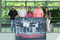 Fünf Personen stehen nebeneinander an einem Metallgeländer in einem hellen Innenraum mit großen Glasfenstern und grünem Baumbestand im Hintergrund. Vor dem Geländer hängt ein großes, dunkles Banner mit einem Gruppenfoto mehrerer Personen in Schwarz‑Weiß. Am unteren Rand des Banners steht in roten Großbuchstaben der Text: „AUSGEBILDET. AUSGELERNT. AUSGESONDERT.“ Die Personen halten das Banner mit beiden Händen; sie tragen unterschiedliche, überwiegend legere Kleidung in dunklen und hellen Farben.