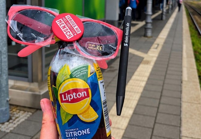 Eine Hand hält eine Lipton-Eisteeflasche mit Zitronengeschmack. An der Flasche sind eine rote Sonnenbrille, ein roter Ansteckbutton mit der Aufschrift „VERGISS NIE HIER REIST EIN MENSCH“ und ein schwarzer Kugelschreiber mit der Aufschrift „5 dein-fonds.de“ befestigt. Im Hintergrund sind ein Bahnsteig und Gleise zu sehen.