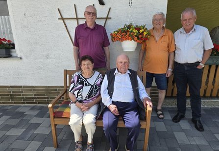 Fünf Personen vor einer hellen Hauswand im Außenbereich. Zwei Personen sitzen auf einer Holzbank im Vordergrund, drei Personen stehen dahinter. Über der Bank hängt ein Blumentopf mit orangefarbenen und gelben Blumen. Links und rechts sind Fensterkästen mit roten Blumen sichtbar.