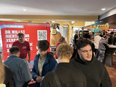 Innenraum mit einem roten Informationsaufsteller der „EVG JUGEND NRW“ im Hintergrund. Auf dem Aufsteller sind ein QR-Code und der teilweise sichtbare Text „DU WILLST NICHTS MEHR VERPASSEN?“ zu sehen. Vor dem Stand stehen mehrere Personen. Rechts im Bild befinden sich weitere Stände, darunter „BAHN BKK“ und „DEVK“, sowie zahlreiche Besucherinnen und Besucher. Im Hintergrund steht eine große dekorative Figur in Tierform.