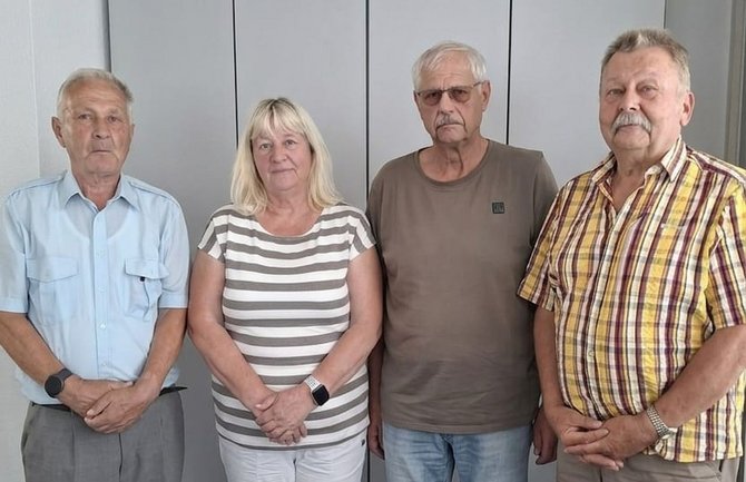 Vier Personen stehen nebeneinander vor einer hellen Wand mit vertikalen Paneelen. Von links nach rechts: eine Person in hellblauem Kurzarmhemd, eine Person mit blondem Haar und gestreiftem Oberteil, eine Person mit Brille und einfarbigem T-Shirt sowie eine Person in kariertem Kurzarmhemd. Alle stehen mit den Händen vor dem Körper und blicken in Richtung Kamera. Der Raum ist hell ausgeleuchtet.