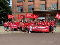 Eine große Gruppe von Personen mit roten EVG-Westen und Fahnen steht vor einem Backsteingebäude und hält ein Banner mit der Aufschrift „WIR STREIKEN“ für ein Gruppenfoto. Vor dem Eingang eines großen Backsteingebäudes hat sich eine Gruppe von rund 35 bis 40 Personen auf dem Gehweg versammelt. Viele tragen rote Warnwesten mit reflektierenden Streifen und dem EVG-Logo. Mehrere Personen halten rote Fahnen der Eisenbahn- und Verkehrsgewerkschaft (EVG) hoch. In der ersten Reihe wird ein großes rotes Banner gehalten. Darauf steht „WIR STREIKEN“ sowie „KEIN ABSCHLUSS OHNE GEWERKSCHAFT“. Einige Personen haben einen Arm erhoben. Vor der Gruppe verläuft eine Straße.