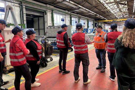 Mehrere Personen mit roten Warnwesten und blauen Kappen stehen in einer großen Werkhalle und hören einer Person in orangefarbener Warnkleidung zu. Die Halle enthält Maschinen, Arbeitsbereiche, Fenster, Hallenschilder und markierte Laufwege auf dem Boden. Auf einigen Warnwesten steht „Besucher“.