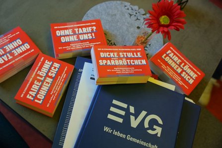 Auf einem Tisch liegen rote Informationskarten und blaue Mappen mit dem Aufdruck „EVG – Wir leben Gemeinschaft“. Auf den Karten stehen unter anderem die Texte „OHNE TARIF? OHNE UNS!“, „DICKE STULLE STATT SPARBRÖTCHEN“ und „FAIRE LÖHNE LOHNEN SICH“. Daneben steht eine rote Blume in einer Vase.