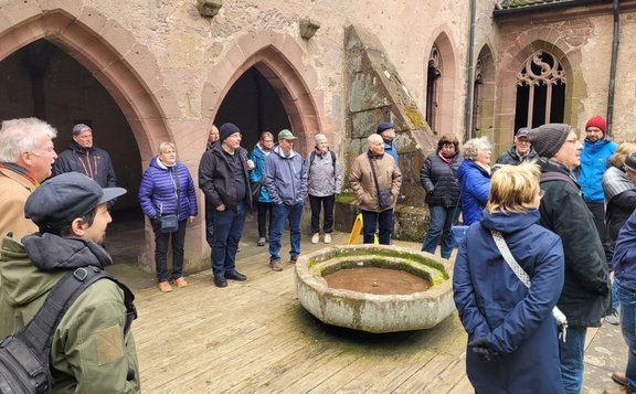 Eine Gruppe von Menschen steht in einem historischen Innenhof vor einer Sandsteinmauer mit spitzbogigen Arkaden. In der Mitte befindet sich ein runder, steinerner Trog mit Wasser. Die Personen tragen Jacken und Mützen und stehen locker verteilt um den Trog. Im Hintergrund sind weitere gotische Fensteröffnungen sichtbar.