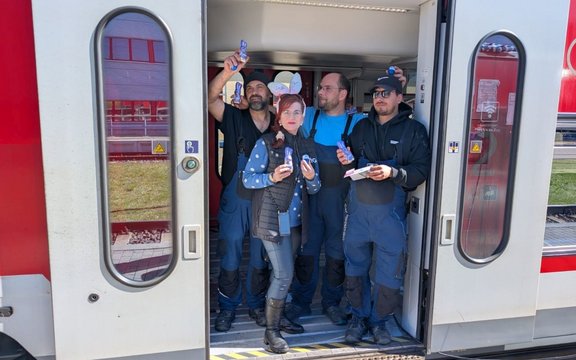 Fünf Personen stehen dicht gedrängt im geöffneten Einstieg eines rot-weißen Zuges an einem Bahnsteig. Die Schiebetüren sind offen, an der Tür sind ein Rollstuhl-Symbol und gelbe Warnaufkleber sichtbar. Die Personen tragen Arbeitskleidung in dunklen Farben; eine Person vorne trägt eine gepunktete Jacke und Stiefel. Mehrere Personen halten kleine Flaschen und verpackte Gegenstände in den Händen, teils hochgehoben. Im Hintergrund ist der Bahnsteig mit Schienen und Gebäudeteilen zu sehen.