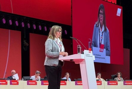 Eine Person steht seitlich hinter einem weißen Rednerpult auf einer Bühne mit rotem Hintergrund. Auf dem Pult sind zwei Mikrofone, ein Glas mit Wasser und ein Schild mit der Aufschrift „DGB“ sowie „23. Ordentlicher Bundeskongress des DGB“. Hinter der Person ist eine große Leinwand zu sehen, die dieselbe Szene in Nahaufnahme zeigt. Im Hintergrund sitzen mehrere Personen an einer langen Tischreihe mit Namensschildern, davor rote Frontblenden. Über der Bühne sind Reihen von runden Bühnenleuchten angebracht.