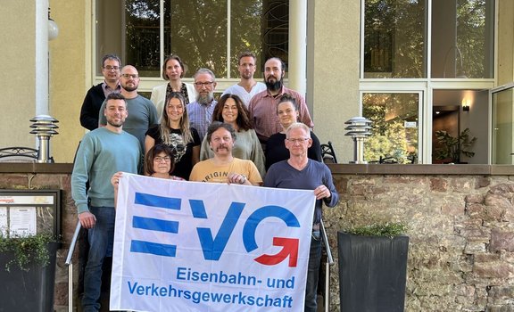 Gruppenfoto vor einem Gebäude mit großen Fenstern und einer Natursteinmauer. Dreizehn Personen stehen auf einer Treppe in mehreren Reihen. Im Vordergrund halten zwei Personen ein weißes Banner mit blau-rotem Logo und dem sichtbaren Text „EVG Eisenbahn- und Verkehrsgewerkschaft“. Links und rechts der Treppe befinden sich Geländer und Leuchten. Durch die Fenster sind Bäume und Teile des Innenraums sichtbar.