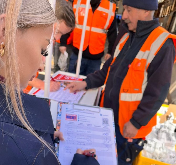 Nahaufnahme eines Tisches, an dem mehrere Personen stehen. Eine Person hält ein Klemmbrett mit einem Formular und schreibt darauf. Eine andere Person zeigt mit dem Finger auf das Papier. Mehrere Personen tragen orangefarbene Warnwesten. Auf dem Tisch liegen weitere Unterlagen und Wasserflaschen.