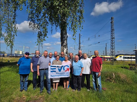 Eine Gruppe von Personen steht im Freien neben Bahngleisen und hält ein Banner der EVG (Eisenbahn- und Verkehrsgewerkschaft). Die Gruppe steht auf einer Wiese unter einem Baum mit hängenden Zweigen. Insgesamt sind mehrere Personen nebeneinander aufgestellt und schauen in Richtung Kamera. In der Mitte halten mehrere Personen ein weißes Banner mit blauem Schriftzug „EVG“ sowie dem Text „Eisenbahn- und Verkehrsgewerkschaft“. Im Hintergrund sind Bahngleise, Oberleitungen und ein stehender Zug zu sehen. Mehrere Strommasten und technische Anlagen entlang der Gleise sind sichtbar. Der Himmel ist blau mit einzelnen weißen Wolken. Die Kleidung der Personen ist überwiegend leger, darunter Hemden, T-Shirts und kurze Hosen.