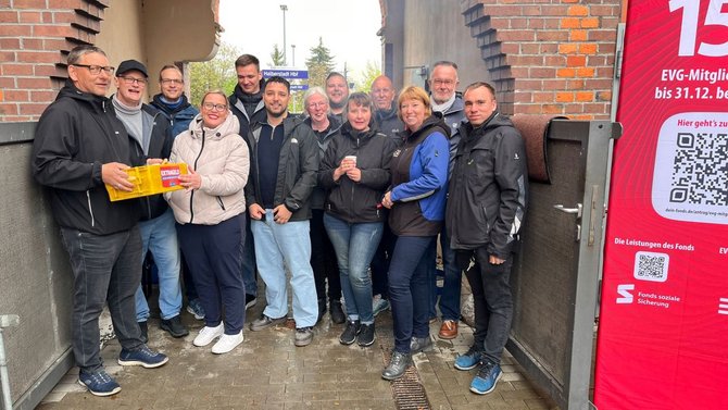 Gruppe von etwa zwölf Personen steht dicht beieinander unter einem gemauerten Durchgang. Zwei Personen halten eine gelbe Kiste mit der Aufschrift „EXTRA“. Rechts daneben hängt ein rotes Banner mit Text, QR-Code und dem Hinweis „EVG-Mitglied“ sowie „Fonds soziale Sicherung“. Im Hintergrund ist ein Bahnhofsschild „Halberstadt Hbf“ sichtbar.