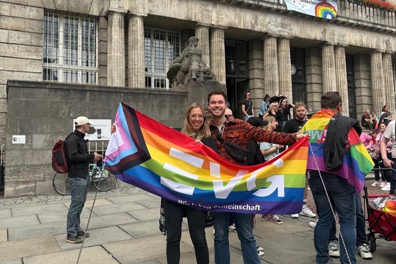 Zwei Personen stehen vor einem großen Gebäude mit Säulen und halten eine bunte Fahne mit dem Schriftzug „ver.di“ in Regenbogenfarben. Im Hintergrund sind weitere Menschen und mehrere Regenbogenflaggen zu sehen. Im Vordergrund stehen zwei Personen auf einer gepflasterten Fläche vor einem monumentalen Gebäude aus Stein mit hohen Säulen und einer Skulptur über dem Eingangsbereich. Die beiden Personen halten gemeinsam eine große ver.di-Fahne, die in den Farben der Regenbogenflagge gestaltet ist und zusätzliche Farbstreifen am linken Rand zeigt. Auf der Fahne ist der Schriftzug „ver.di“ groß und weiß abgebildet.