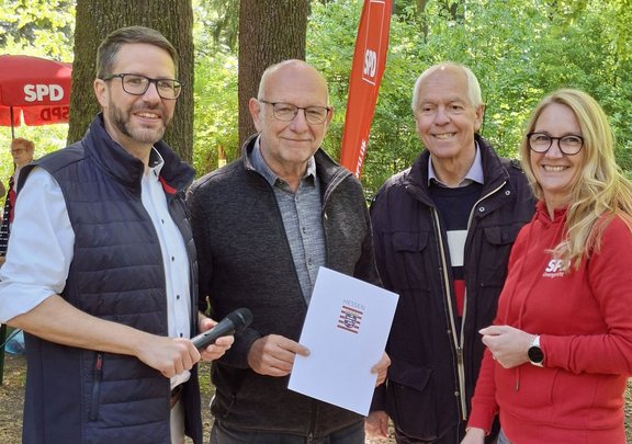 Im Freien stehen vier erwachsene Personen dicht nebeneinander vor Bäumen. Drei Personen tragen dunkle Jacken, eine Person trägt einen roten Kapuzenpullover mit weißem „SPD“-Aufdruck. Eine Person hält ein schwarzes Handmikrofon, eine andere hält ein weißes Blatt Papier mit dem Aufdruck „HESSEN“ und einem Wappen. Im Hintergrund sind rote Fahnen und ein roter Sonnenschirm mit der Aufschrift „SPD“ zu sehen.