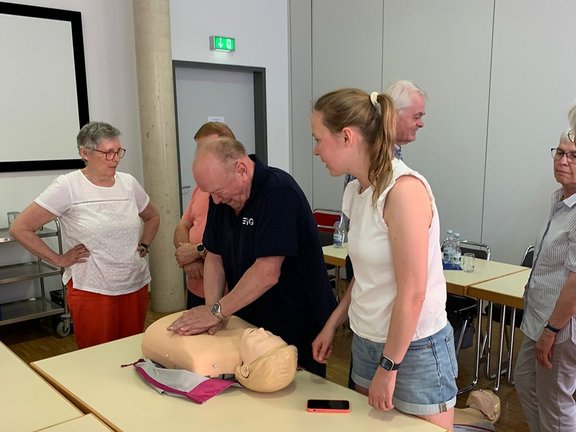 Mehrere Personen üben an einer Wiederbelebungs‑Puppe die Herz‑Lungen‑Wiederbelebung in einem Seminarraum. Im Vordergrund liegt eine Übungspuppe auf einem Tisch. Eine Person drückt mit beiden Händen auf den Brustbereich der Puppe und führt damit die Herzdruckmassage aus. Daneben stehen mehrere weitere Personen und beobachten die Übung. Auf dem Tisch liegt eine geöffnete Tasche, in der Nähe befindet sich ein Smartphone.