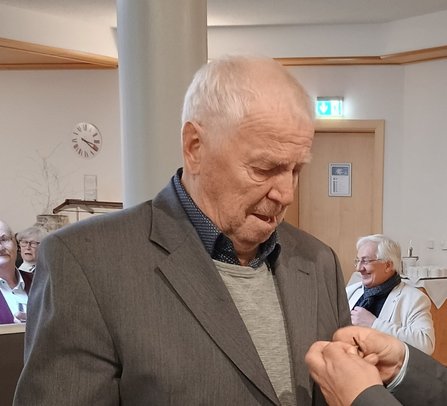 Eine Person in grauem Sakko über hellem Pullover steht in einem Innenraum und blickt nach unten auf die eigenen Hände. Rechts im Vordergrund sind die Hände einer weiteren Person sichtbar. Im Hintergrund befinden sich mehrere sitzende Personen, eine Uhr an der Wand, eine Tür und ein beleuchtetes Notausgangsschild.