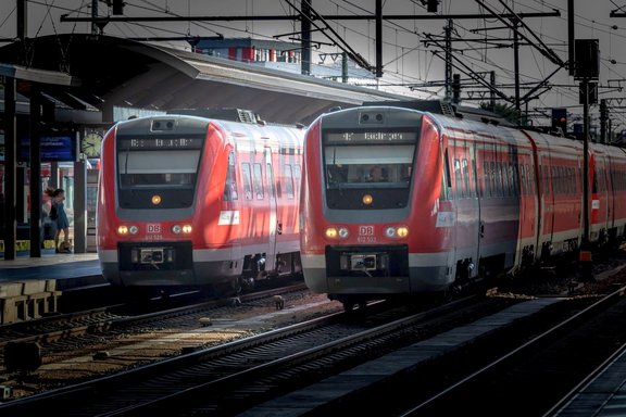 Zwei rote Regionalzüge der Deutschen Bahn stehen nebeneinander auf parallelen Gleisen in einem Bahnhof. Über den Zügen verlaufen zahlreiche Oberleitungen und Strommasten. Links ist ein Bahnsteig mit Überdachung, einer Uhr und einer einzelnen Person zu sehen. Die Fronten der Züge sind sichtbar, an einem Zug ist die Nummer „612 529“, am anderen „612 533“ erkennbar. Die Szene ist von hellem Sonnenlicht und starken Schatten geprägt.
