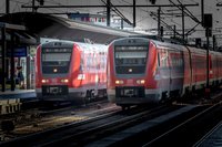 Zwei rote Regionalzüge der Deutschen Bahn stehen nebeneinander auf parallelen Gleisen in einem Bahnhof. Über den Zügen verlaufen zahlreiche Oberleitungen und Strommasten. Links ist ein Bahnsteig mit Überdachung, einer Uhr und einer einzelnen Person zu sehen. Die Fronten der Züge sind sichtbar, an einem Zug ist die Nummer „612 529“, am anderen „612 533“ erkennbar. Die Szene ist von hellem Sonnenlicht und starken Schatten geprägt.