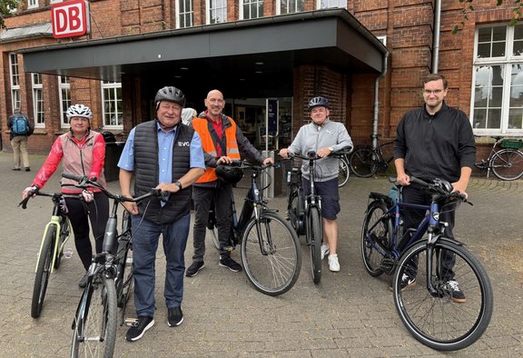 Fünf Personen mit Fahrrädern stehen vor dem Eingang eines Backsteingebäudes mit einem gut sichtbaren DB-Logo. Mehrere Personen tragen Fahrradhelme. Im Hintergrund sind Fenster, abgestellte Fahrräder und weitere Personen zu sehen.