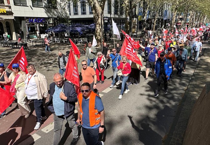 Auf einer breiten Straße mit Bäumen und Cafés gehen viele Menschen in einer dichten Gruppe in eine Richtung. Mehrere Personen tragen rote Fahnen mit weißem Schriftzug, unter anderem „Eisenbahn- und Verkehrsgewerkschaft“ und „EVG“. Einige tragen orangefarbene Warnwesten mit der Aufschrift „EVG“. Im Hintergrund sind Ladenfassaden, Außengastronomie, Fahrräder und weitere Fahnen zu sehen.