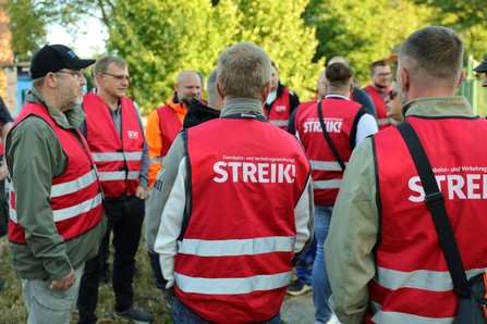 Mehrere Personen in roten Warnwesten stehen im Freien in einem Kreis zusammen. Auf den Westen steht auf der Rückseite „STREIK!“ sowie „Eisenbahn- und Verkehrsgewerkschaft“. Im Hintergrund sind Bäume und weitere Personen sichtbar.