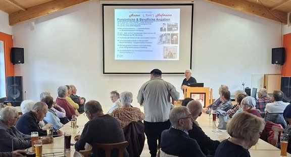 Großer Veranstaltungsraum mit Holzdecke und weißer Frontwand. Mehrere Personen sitzen dicht nebeneinander an Tischen und Stühlen. Auf den Tischen stehen Gläser, Flaschen und Unterlagen. Vorne steht eine Person hinter einem Rednerpult mit Laptop; darüber ist eine Projektionsleinwand. Auf der Folie ist lesbarer Text zu sehen: „Persönliche & Berufliche Angaben“ sowie der Name „Anton („Toni“) Hoffmann“ und mehrere Stichpunkte in Aufzählungsform, ergänzt durch kleine Fotos rechts. Links und rechts der Leinwand stehen Lautsprecherboxen.