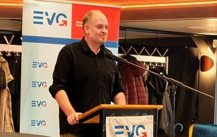 Eine Person steht an einem Rednerpult mit einem Mikrofon. Auf dem Pult und auf einem Roll-up im Hintergrund ist mehrfach das Logo „EVG“ zu sehen. Hinter dem Pult befinden sich Garderobenständer mit aufgehängten Jacken. Der Raum ist mit warmen Lichtleisten an der Decke beleuchtet.