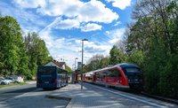 Bahnhaltestelle im Freien mit gepflastertem Bahnsteig: Links steht ein blauer Linienbus an der Haltestelle, rechts hält ein roter Regionalzug auf den Gleisen. In der Mitte des Bahnsteigs stehen mehrere schlanke Laternenmasten und ein Haltestellenschild. Umgeben ist die Haltestelle von grünen Bäumen und Sträuchern; im Hintergrund sind Wohnhäuser zu sehen. Über der Szene ein blauer Himmel mit weißen Wolken.