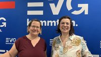 Zwei Personen stehen vor derselben blauen Fotowand mit mehrfach sichtbarem weißen Schriftzug „EVG kommt an.“. Die linke Person trägt ein bordeauxfarbenes Oberteil und eine Brille. Die rechte Person trägt eine helle Bluse mit floralem Muster und eine Brille. Beide sind von der Hüfte aufwärts zu sehen.