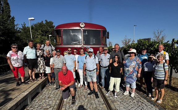 Gruppe von etwa 20 Personen steht und kniet auf Gleisen vor der Front eines roten Schienenfahrzeugs. Links befindet sich ein Bahnsteig mit mehreren Laternen, im Hintergrund Bäume. Rechts sind Pflanzenreihen und weitere Laternen zu sehen. Klarer blauer Himmel und starke Sonneneinstrahlung.