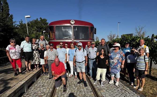 Gruppe von etwa 20 Personen steht und kniet auf Gleisen vor der Front eines roten Schienenfahrzeugs. Links befindet sich ein Bahnsteig mit mehreren Laternen, im Hintergrund Bäume. Rechts sind Pflanzenreihen und weitere Laternen zu sehen. Klarer blauer Himmel und starke Sonneneinstrahlung.