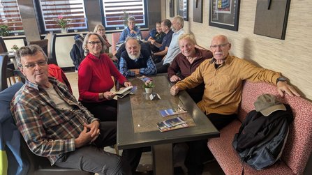 Mehrere Personen sitzen an Tischen in einem Café oder Restaurant. Im Vordergrund sitzen fünf Personen an einem rechteckigen Tisch mit Prospekten und einer kleinen Topfpflanze. Im Hintergrund befinden sich weitere Personen an einem zweiten Tisch. An der Wand hängen gerahmte Bilder, dahinter sind Fenster mit gestreiften Jalousien.