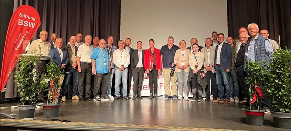Gruppenfoto von etwa zwanzig bis fünfundzwanzig Personen auf einer Bühne vor einer Leinwand, links steht ein roter Aufsteller mit der Aufschrift „Stiftung BSW Ortsstelle Frohnberg“. Die Personen stehen in einer Reihe nebeneinander auf einer erhöhten Bühne und schauen in Richtung Kamera. Hinter der Gruppe hängt eine große helle Leinwand, die von dunklen Vorhängen eingerahmt ist. Links neben der Gruppe steht ein roter, tropfenförmiger Banner mit weißer Schrift. Auf der Bühne sind mehrere grüne Pflanzen in Töpfen platziert, besonders im Vordergrund links und rechts. Einige Personen tragen Namensschilder um den Hals, andere halten Jacken oder Taschen in den Händen. Mehrere Personen tragen helle Hemden oder Jacken, einzelne sind farblich hervorgehoben, zum Beispiel durch einen roten Blazer.