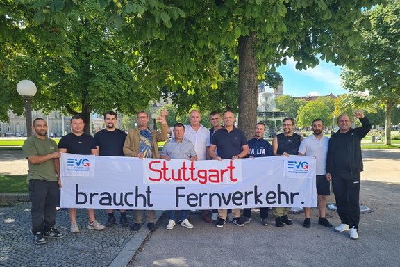 Mehrere Personen stehen in einem Park hinter einem Transparent mit der Aufschrift „Stuttgart braucht Fernverkehr“. Die Gruppe hat sich auf einem Weg unter großen Bäumen aufgestellt und hält gemeinsam ein langes weißes Transparent. Auf dem Transparent steht in großen Buchstaben „Stuttgart braucht Fernverkehr“. An beiden Seiten sind die Logos der EVG zu sehen. Einige Personen haben einen Arm angehoben.