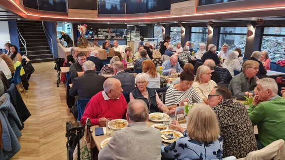 Großer Gastraum mit zahlreichen besetzten Tischen. Viele Personen sitzen beim Essen und unterhalten sich. Im Hintergrund befinden sich eine Theke mit Regalen, große Fenster und eine Treppe. Auf mehreren Tischen stehen Teller, Gläser, Getränkeflaschen und kleine Blumenvasen. Die Decke ist mit umlaufenden Lichtleisten beleuchtet.