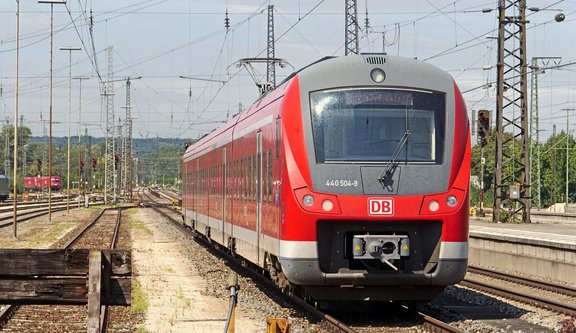 Roter Regionalzug mit grauer Front und sichtbarem DB‑Logo steht auf mehreren Gleisen in einem Bahnhofs- oder Abstellbereich; im Vordergrund verläuft ein Gleis, im Hintergrund sind weitere Gleise, Oberleitungen, Masten, Signale und Bahnanlagen zu sehen, umgeben von Bäumen und leicht hügeliger Landschaft unter hellem Himmel.