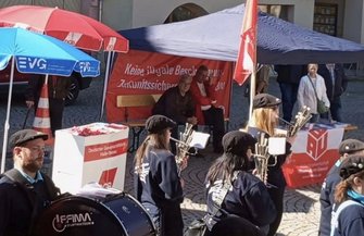 Mehrere Personen mit Musikinstrumenten, darunter Trommeln und Blasinstrumente, stehen vor mehreren Infoständen. Im Hintergrund sind rote und blaue Pavillons mit EVG‑ und DGB‑Logos sowie ein rotes Banner mit weißer Schrift zu sehen. Auf einem weißen Stand ist „Deutscher Gewerkschaftsbund“ lesbar. Der Platz ist gepflastert.