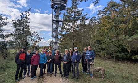 Eine Gruppe von Personen steht auf einem Weg vor einem hohen Aussichtsturm mit spiralförmig verlaufender Außentreppe. Rechts im Vordergrund steht ein Hund an einer Leine. Um den Turm befinden sich Bäume, im Hintergrund sind Wiesen sowie ein teilweise bewölkter Himmel zu sehen.