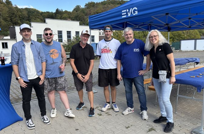 Sechs Personen stehen auf einer gepflasterten Fläche vor einem blauen Pavillon mit dem sichtbaren Schriftzug „EVG“. Im Hintergrund befinden sich ein helles Gebäude, bewaldete Hänge und mehrere Biertischgarnituren. Die Personen tragen Freizeitkleidung, darunter Kappen, T-Shirts, Hemden, Shorts und Jeans. Auf einem Stehtisch links steht eine Getränkeflasche.