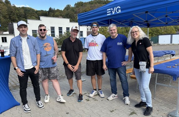 Sechs Personen stehen auf einer gepflasterten Fläche vor einem blauen Pavillon mit dem sichtbaren Schriftzug „EVG“. Im Hintergrund befinden sich ein helles Gebäude, bewaldete Hänge und mehrere Biertischgarnituren. Die Personen tragen Freizeitkleidung, darunter Kappen, T-Shirts, Hemden, Shorts und Jeans. Auf einem Stehtisch links steht eine Getränkeflasche.