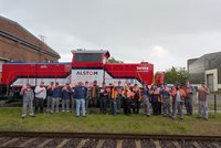 Gruppenfoto im Freien: Etwa 25 Personen stehen nebeneinander auf Gras vor einem roten Schienenfahrzeug mit blau-weißen Streifen. Auf dem Fahrzeug sind die gut lesbaren Schriftzüge „ALSTOM“, „Lokomotiven Service“, „Zürich“ und „Service ist unser Antrieb“ angebracht. Die Personen tragen überwiegend Arbeitskleidung in Grau und Blau, einige tragen orangefarbene Warnwesten und Schutzhelme. Viele halten kleine rote Schilder mit weißem Symbol oder Schrift in die Kamera. Im Vordergrund verlaufen Eisenbahnschienen. Im Hintergrund sind Backstein- und Industriegebäude sowie ein bewölkter Himmel zu sehen.